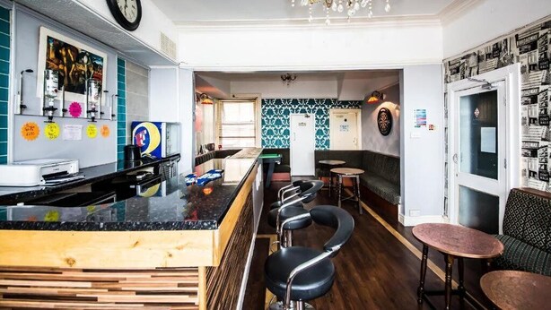 Zona de bar con mostrador de madera, taburetes negros y un candelabro. Hay un reloj en la pared y un cartel con un mensaje.