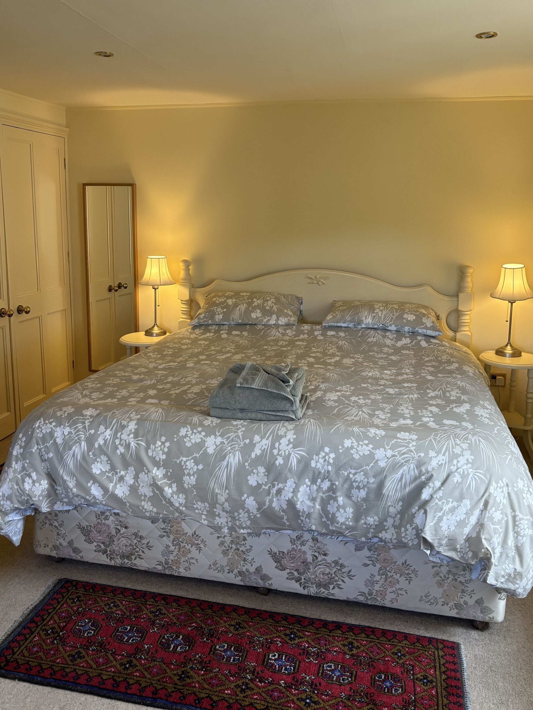 A bed with floral bedding, two bedside lamps, a red patterned rug, and a wardrobe with a door.