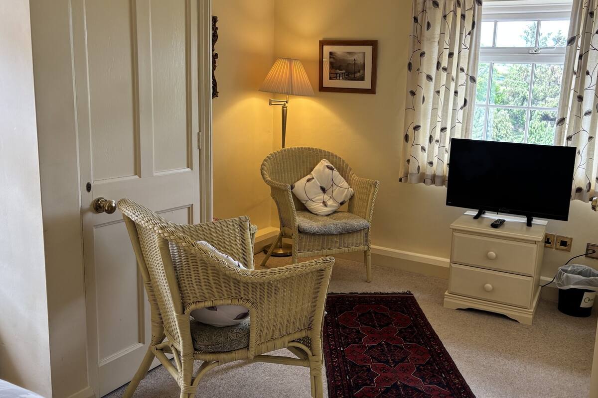 A room with a wicker chair, a television on a stand, a lamp, and a window with curtains.