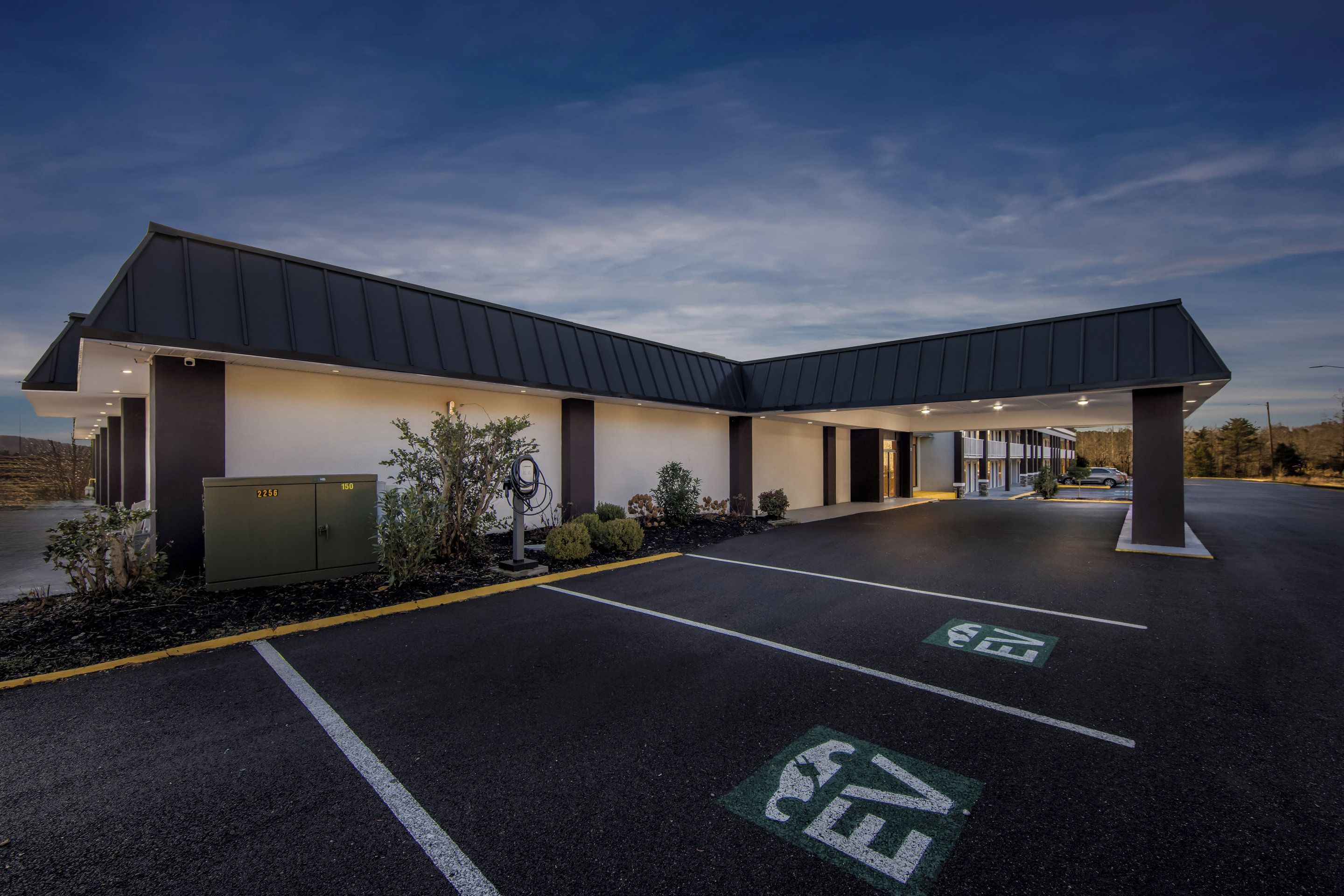 Un edificio moderno con zona de estacionamiento techada, estaciones de carga para vehículos eléctricos señalizadas y un cielo despejado.