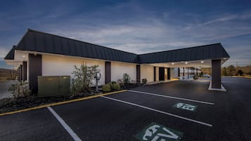 Un edificio moderno con zona de estacionamiento techada, estaciones de carga para vehículos eléctricos señalizadas y un cielo despejado.