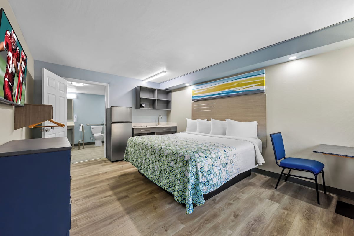 A hotel room with a bed, a desk, a chair, a small kitchenette, and a bathroom.