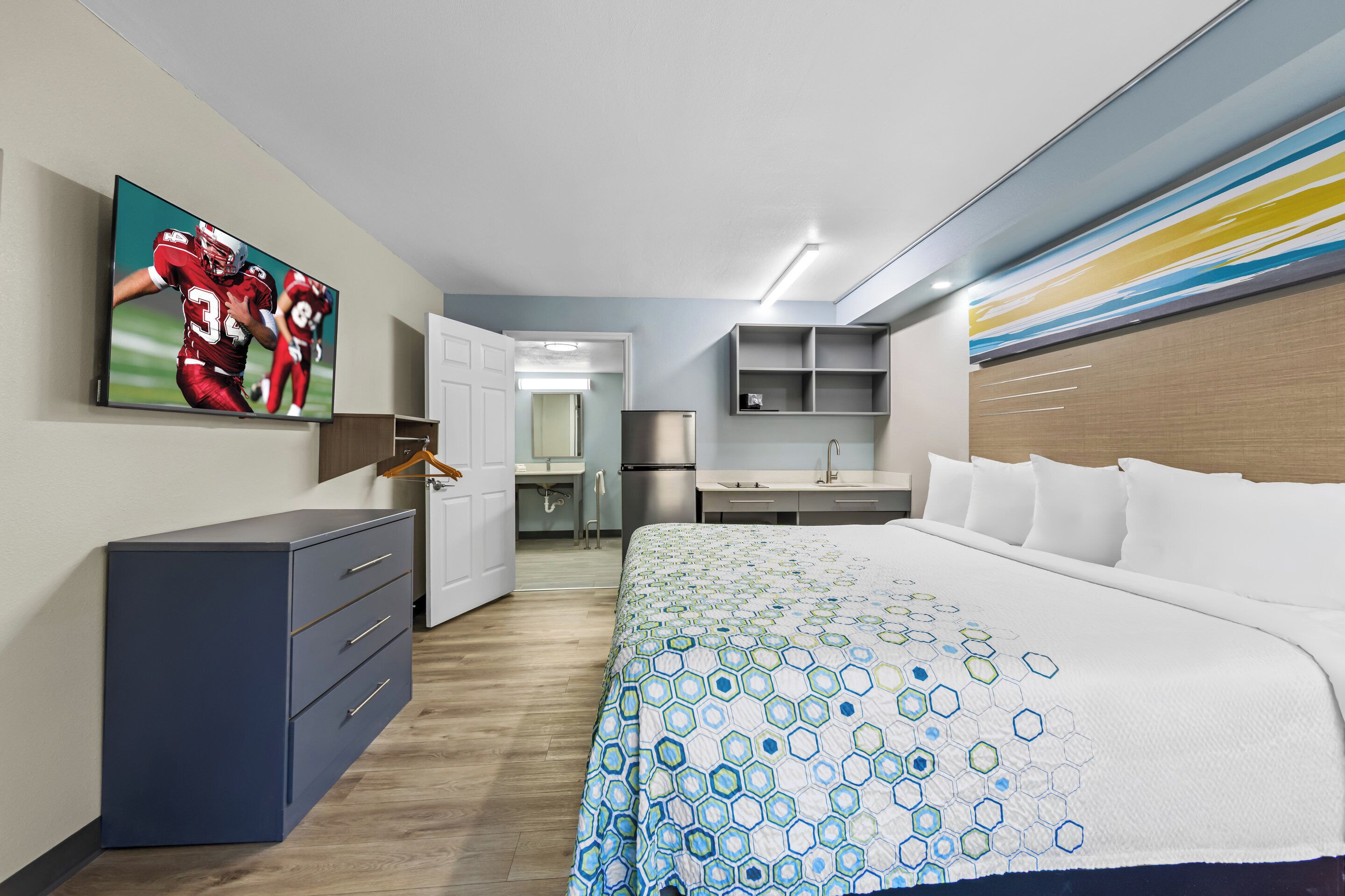A modern hotel room with a large bed, a desk, a wardrobe, and a television displaying a sports game.
