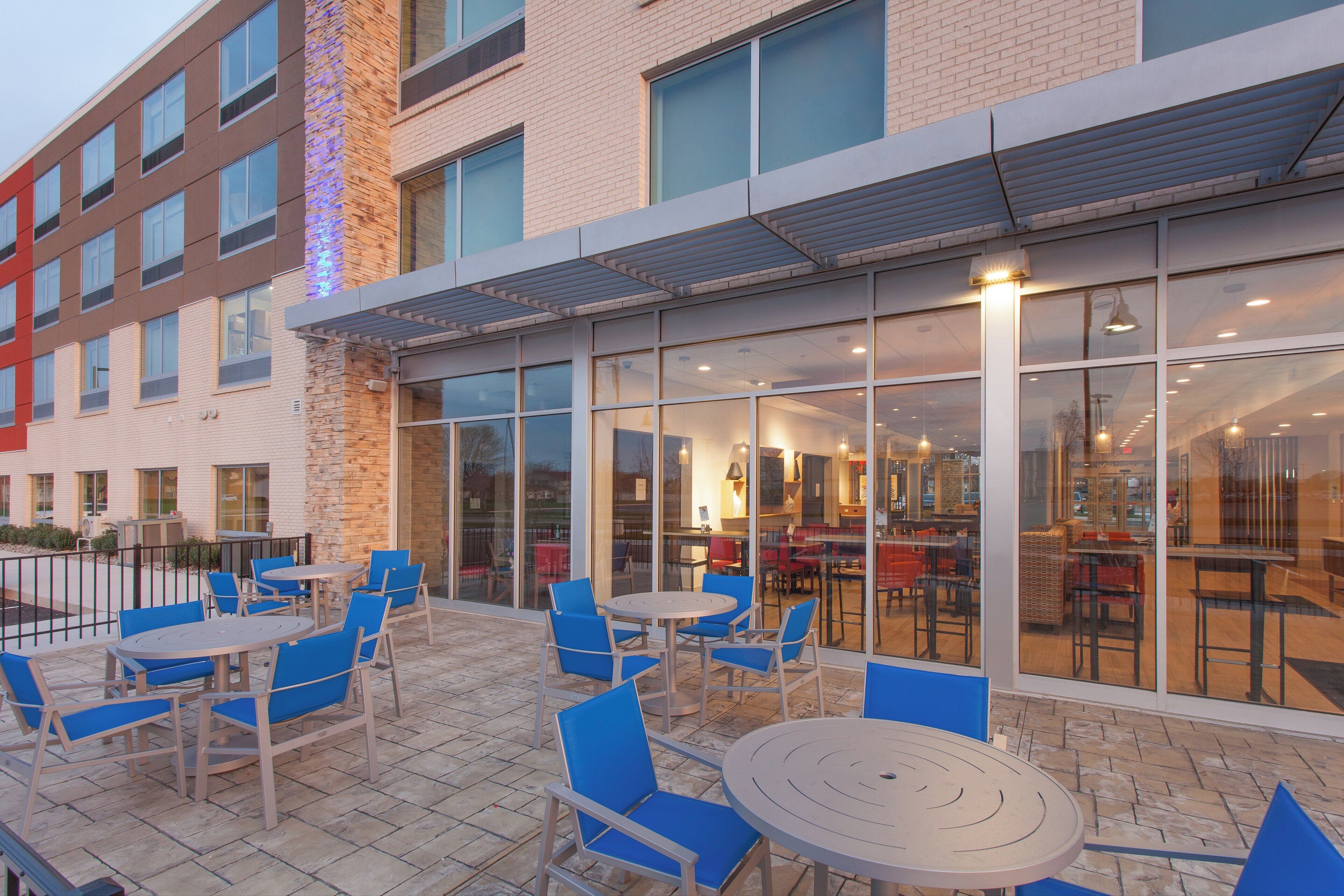 A building with a glass facade, outdoor seating area with tables and chairs, and a brick wall.