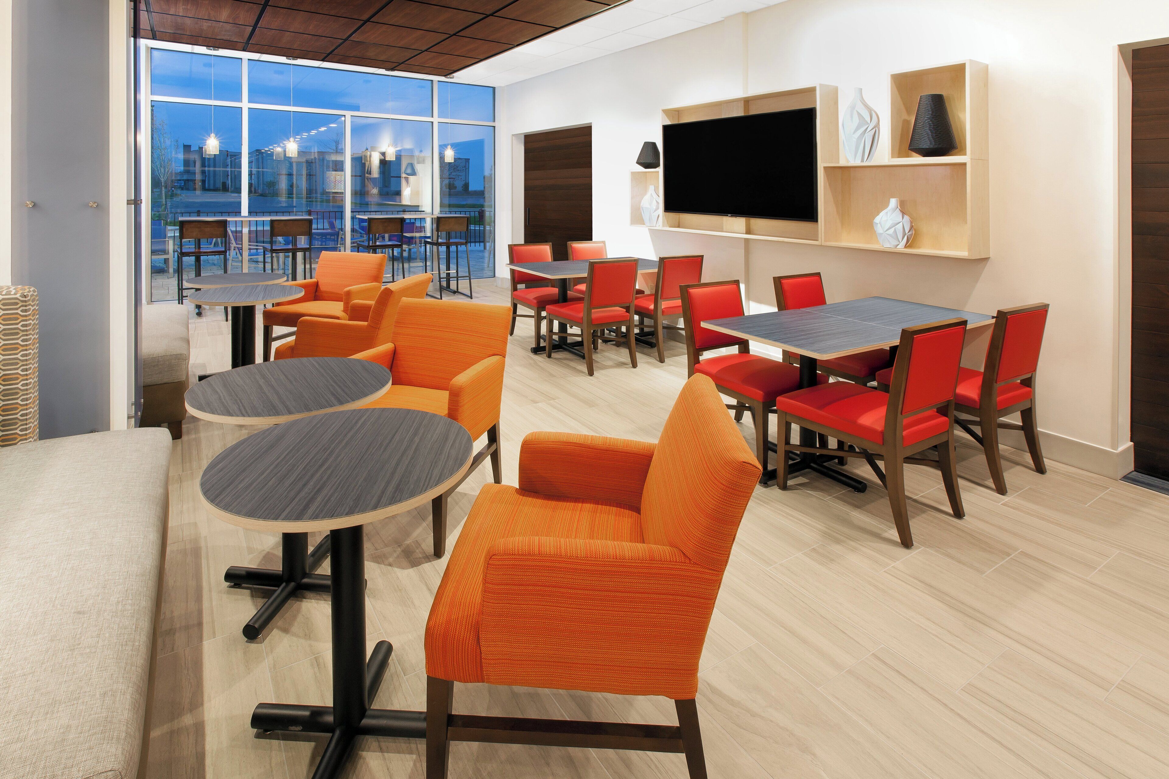 A modern dining area with round tables and orange chairs, a flat-screen TV on the wall, and large windows offering a view of the city.