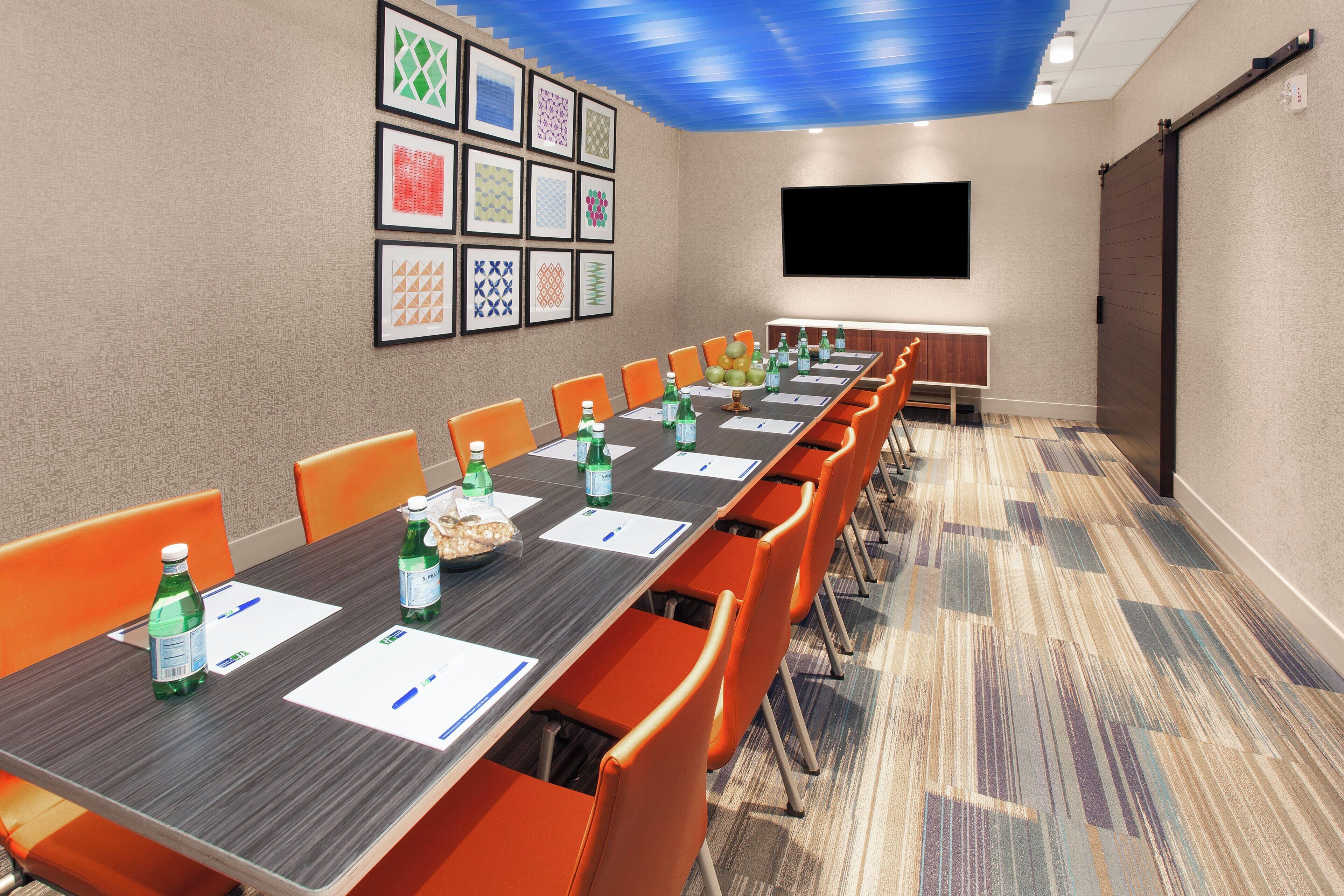 A conference room with a long table, orange chairs, water bottles, and a projector screen.