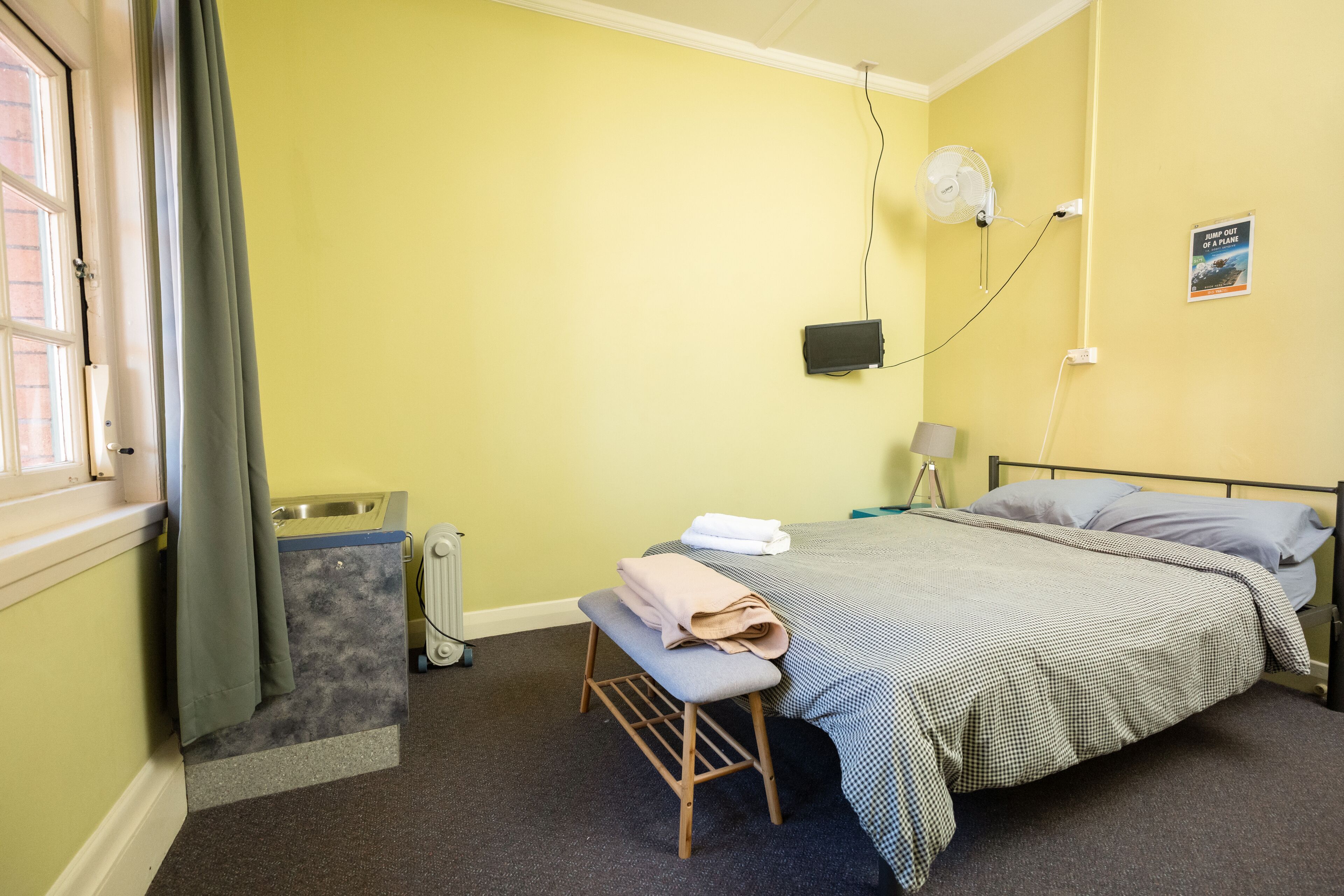 Une pièce comprenant un lit, un banc, un radiateur, une fenêtre avec des rideaux, un ventilateur fixé au mur et une affiche accrochée au mur.