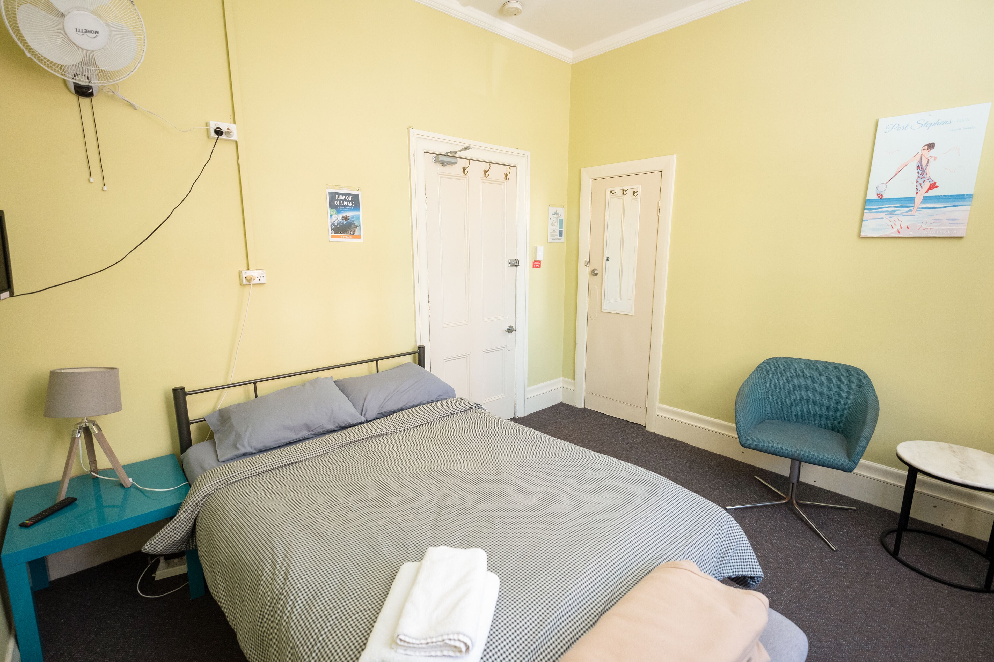 A hotel room with a bed, a chair, a small table, a fan, and a wall-mounted picture.