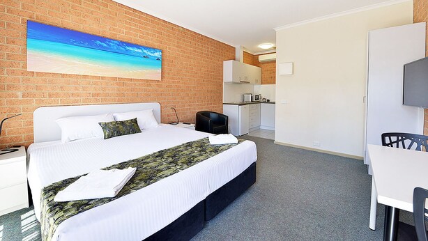 A hotel room with a bed, a desk, a chair, a TV, and a kitchen area. - Cranbourne Motor Inn (Cranbourne)