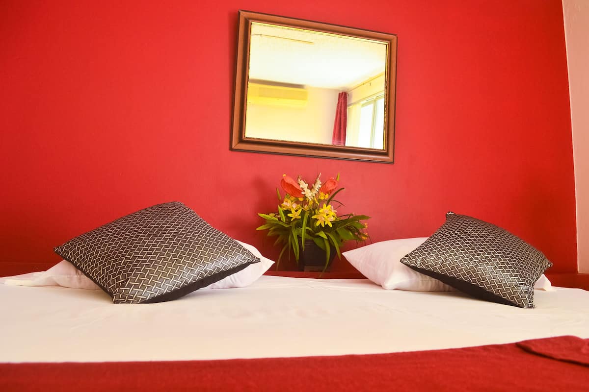 Une chambre d’hôtel avec un mur rouge, un lit avec une literie blanche, deux coussins à motifs et un vase rempli de fleurs jaunes.