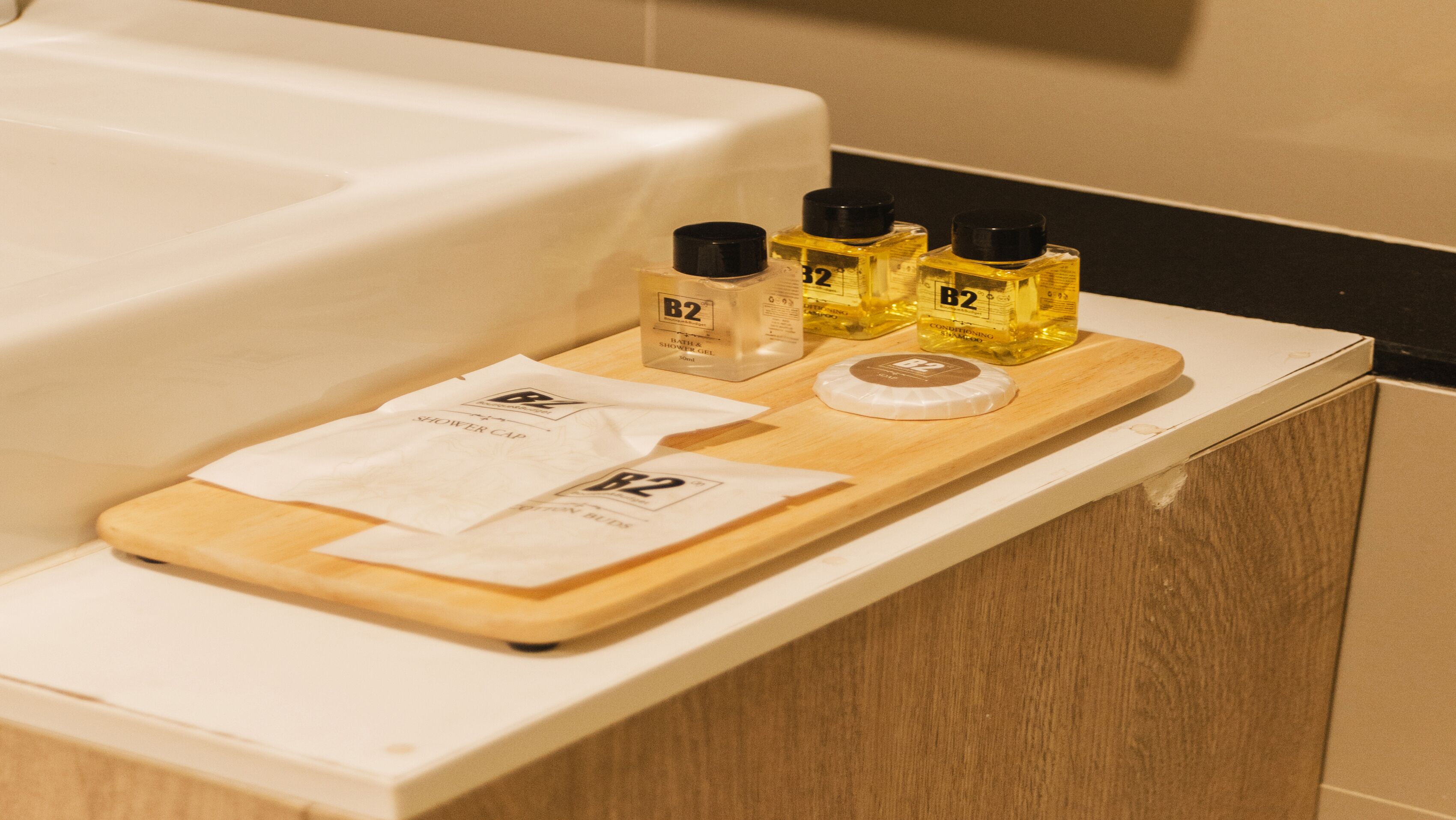 Ein Badezimmer-Waschtisch mit einer Holztasse, die eine Duschhaube, eine Flasche B2 Bath & Shower Gel und zwei Flaschen B2 Conditioner Shampoos enthält.