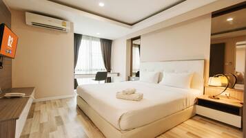 A hotel room with a large bed, a desk, a chair, a TV, and an air conditioning unit.