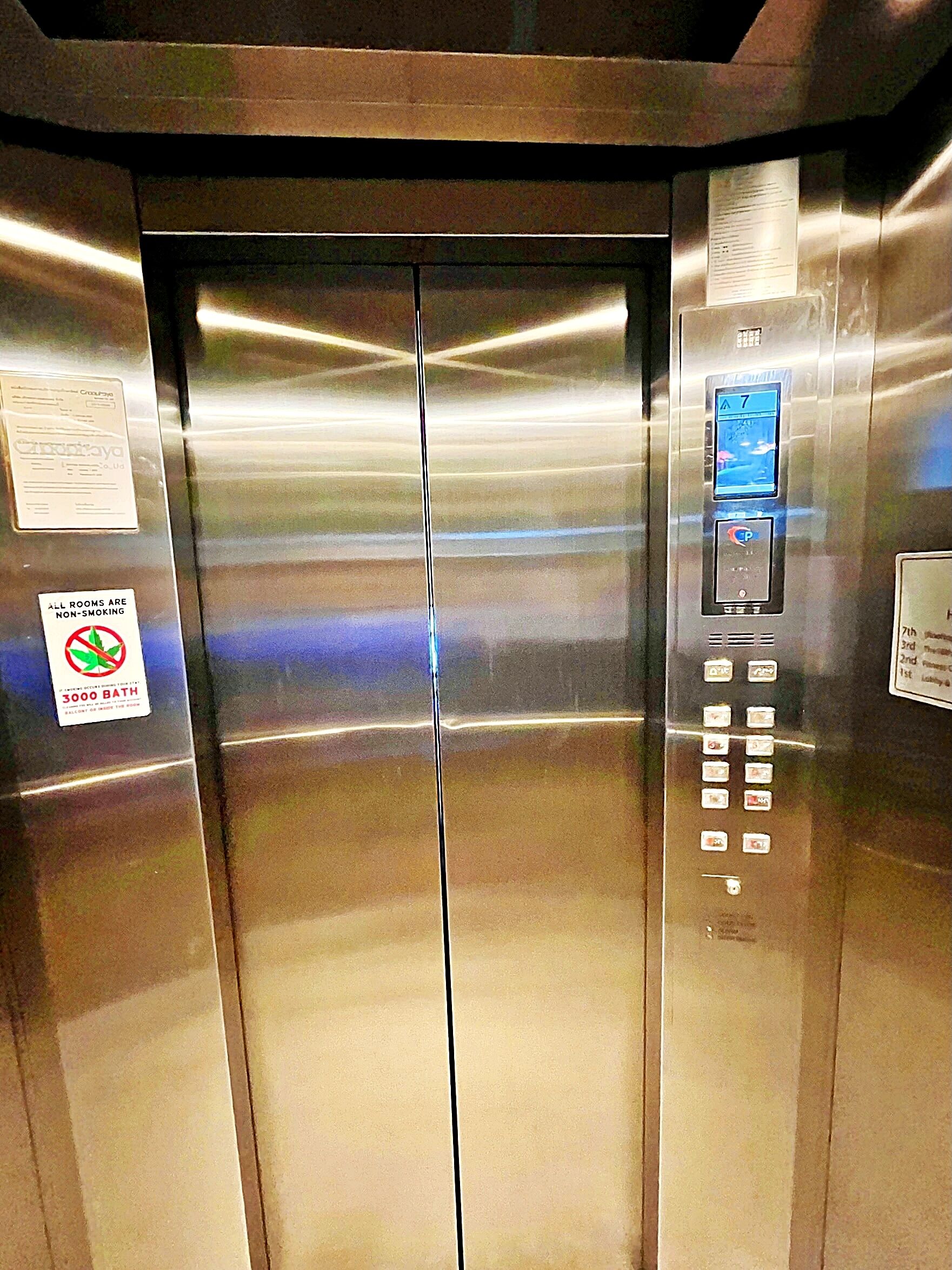Interior d'un ascensor amb acabat metàl·lic, un panell de control amb botons i un pantalla digital.