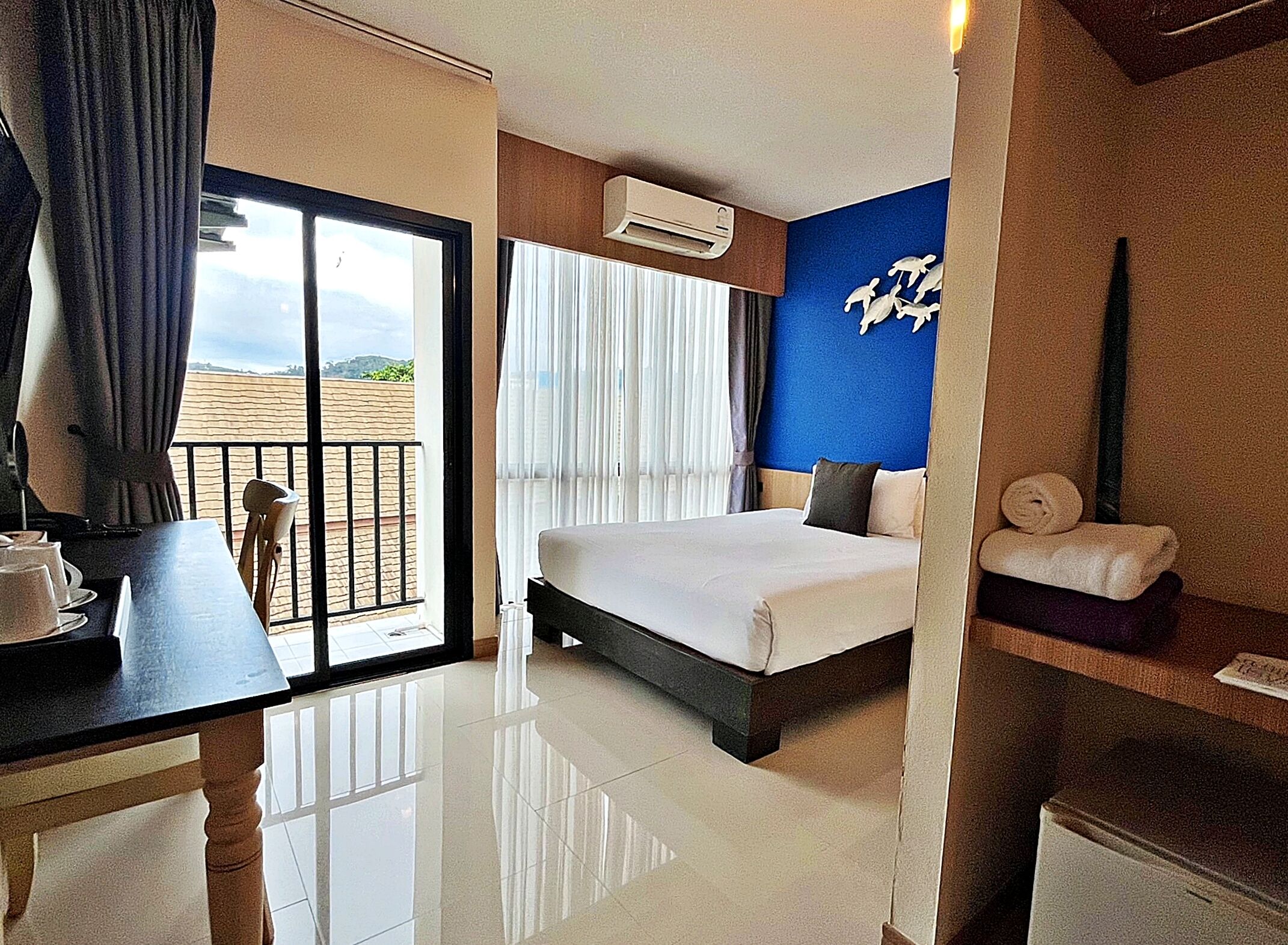 A hotel room with a bed, a desk with a chair, a window with curtains, a wall-mounted air conditioner, and a towel rack with towels.