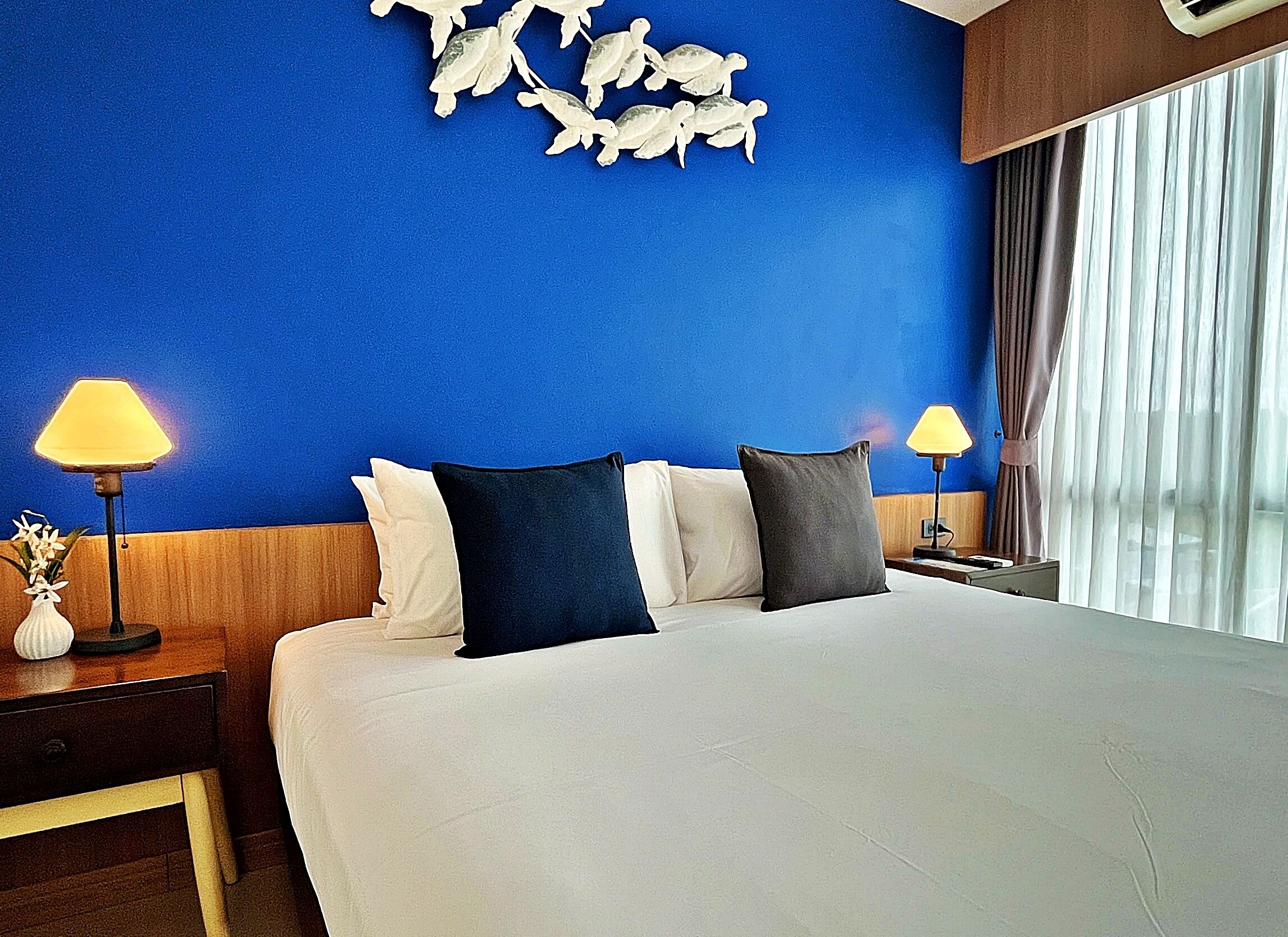 A hotel room with a large bed, two bedside lamps, a nightstand with a vase of flowers, and a blue wall with a decorative wall art piece.