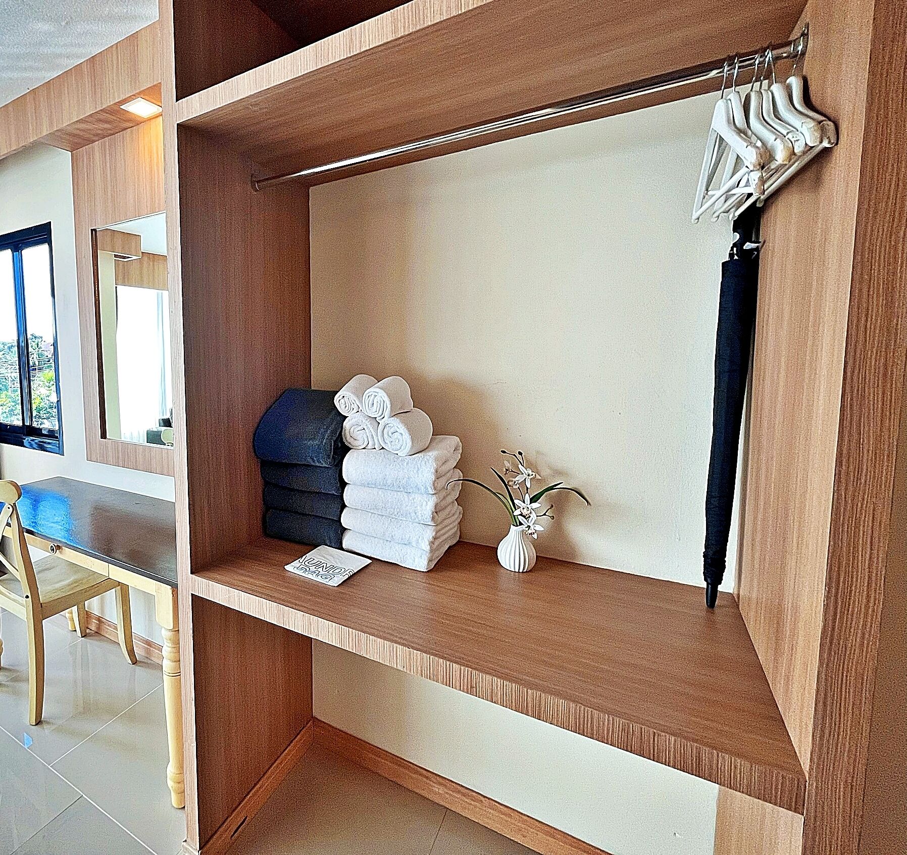 A hotel room with a wooden vanity, a mirror, a towel rack, and a small vase with flowers.