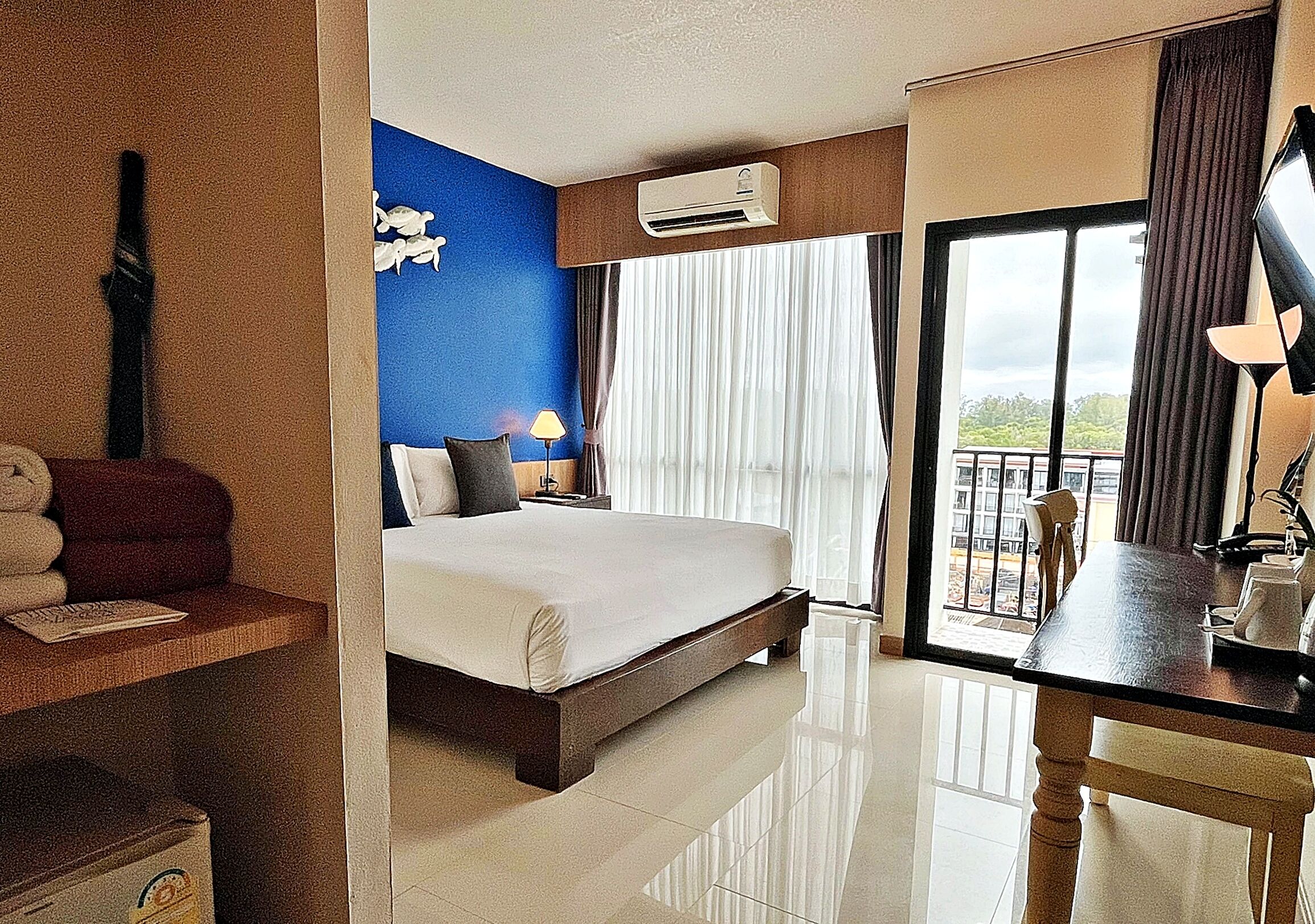 A hotel room with a bed, a desk, a chair, a lamp, an air conditioner, and a window with curtains.