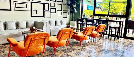 A modern living room with a grey sofa, orange chairs, patterned floor tiles, and a large window with a view of greenery.