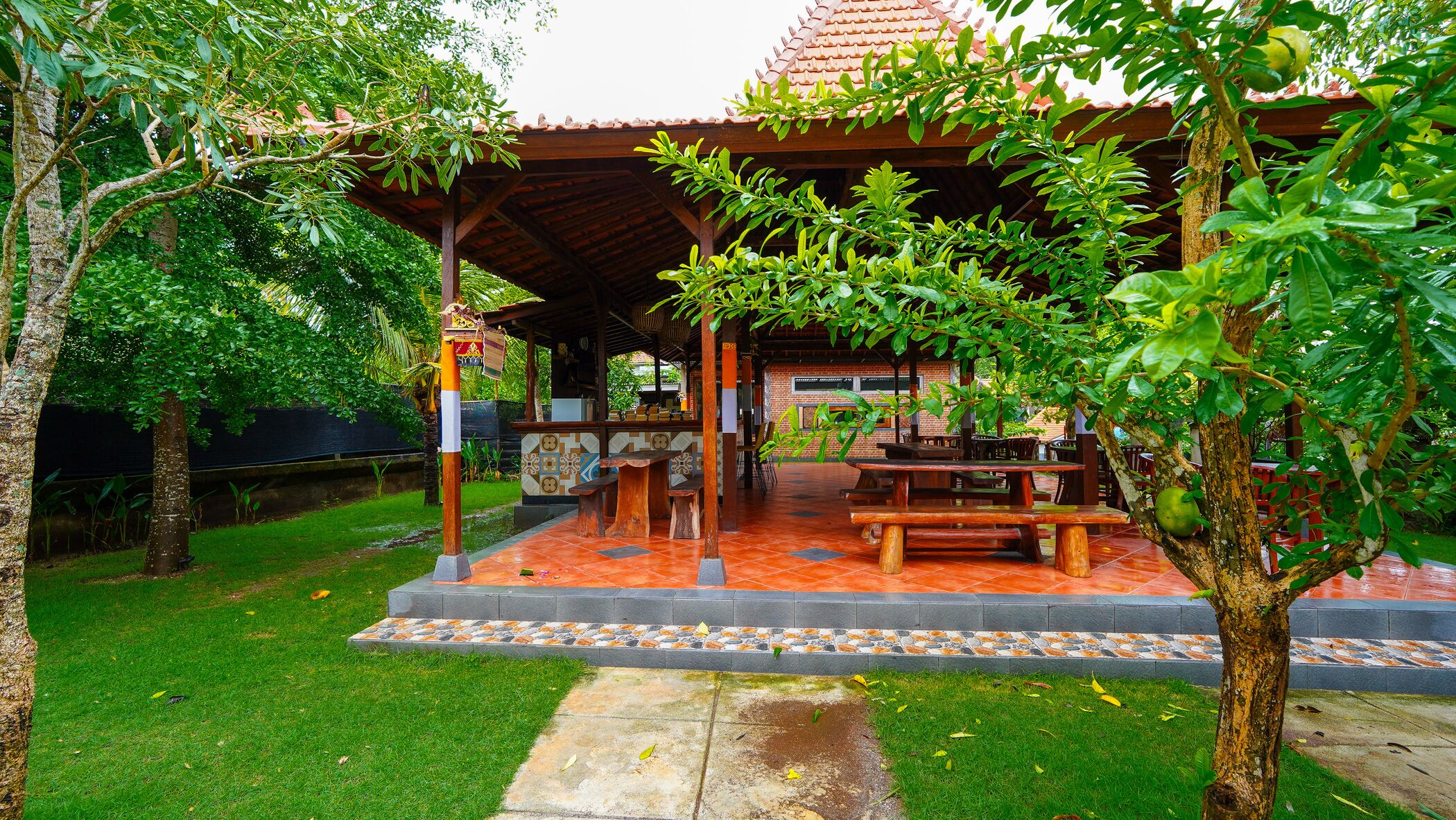 A covered outdoor seating area with wooden tables and benches, surrounded by lush greenery and trees.