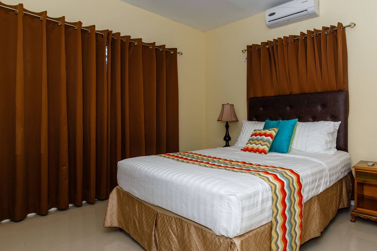 Une chambre à coucher avec un grand lit, des rideaux marron, une tête de lit, une lampe de chevet et une table de chevet en bois.