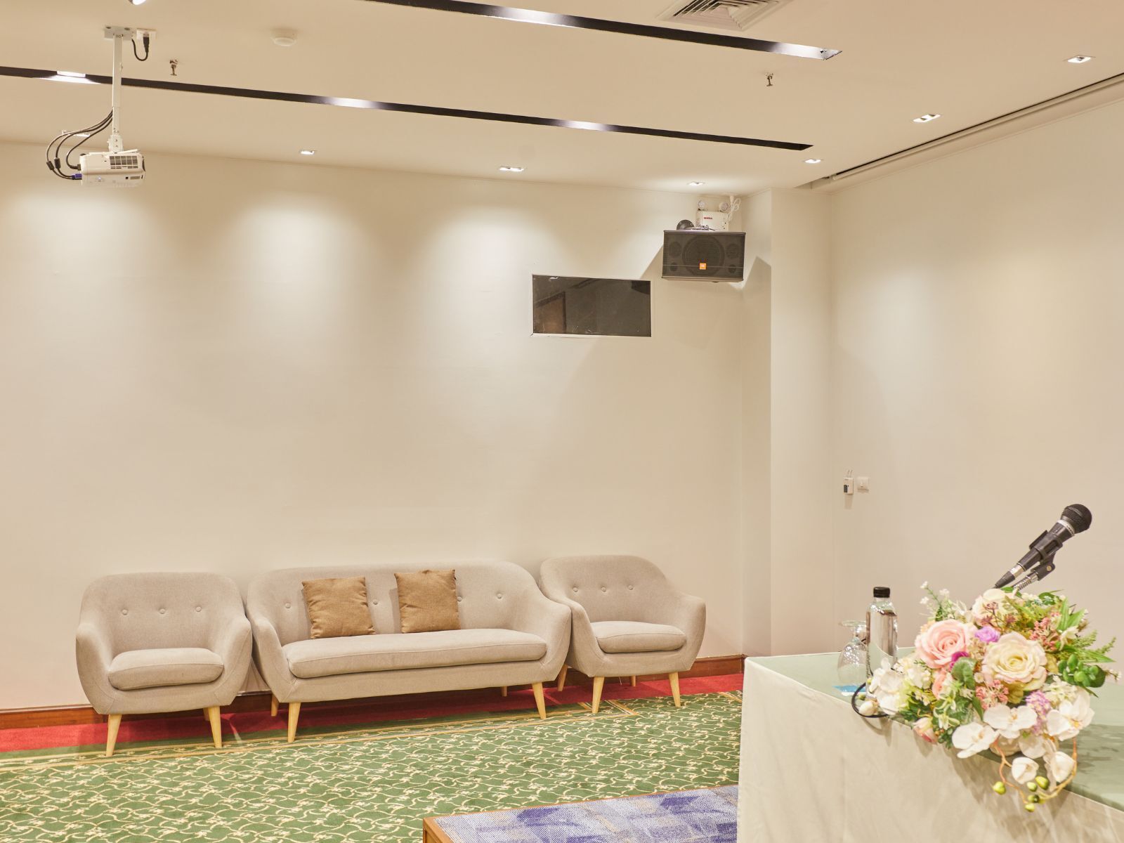 Una sala de conferencias con proyector, una pantalla grande, tres sofás, una mesa con un micrófono y flores, y una alfombra estampada.