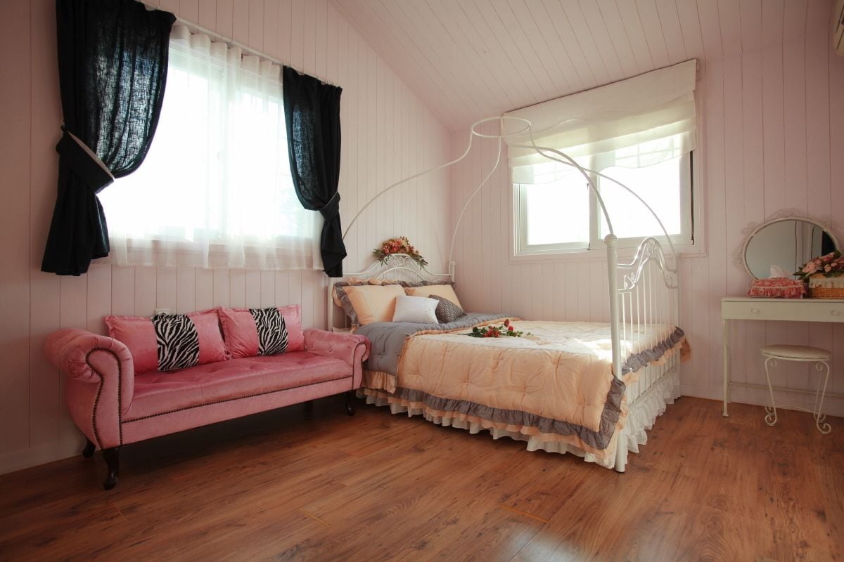 A bedroom with a pink sofa, a bed with a canopy, a desk with a mirror, and a window with curtains.