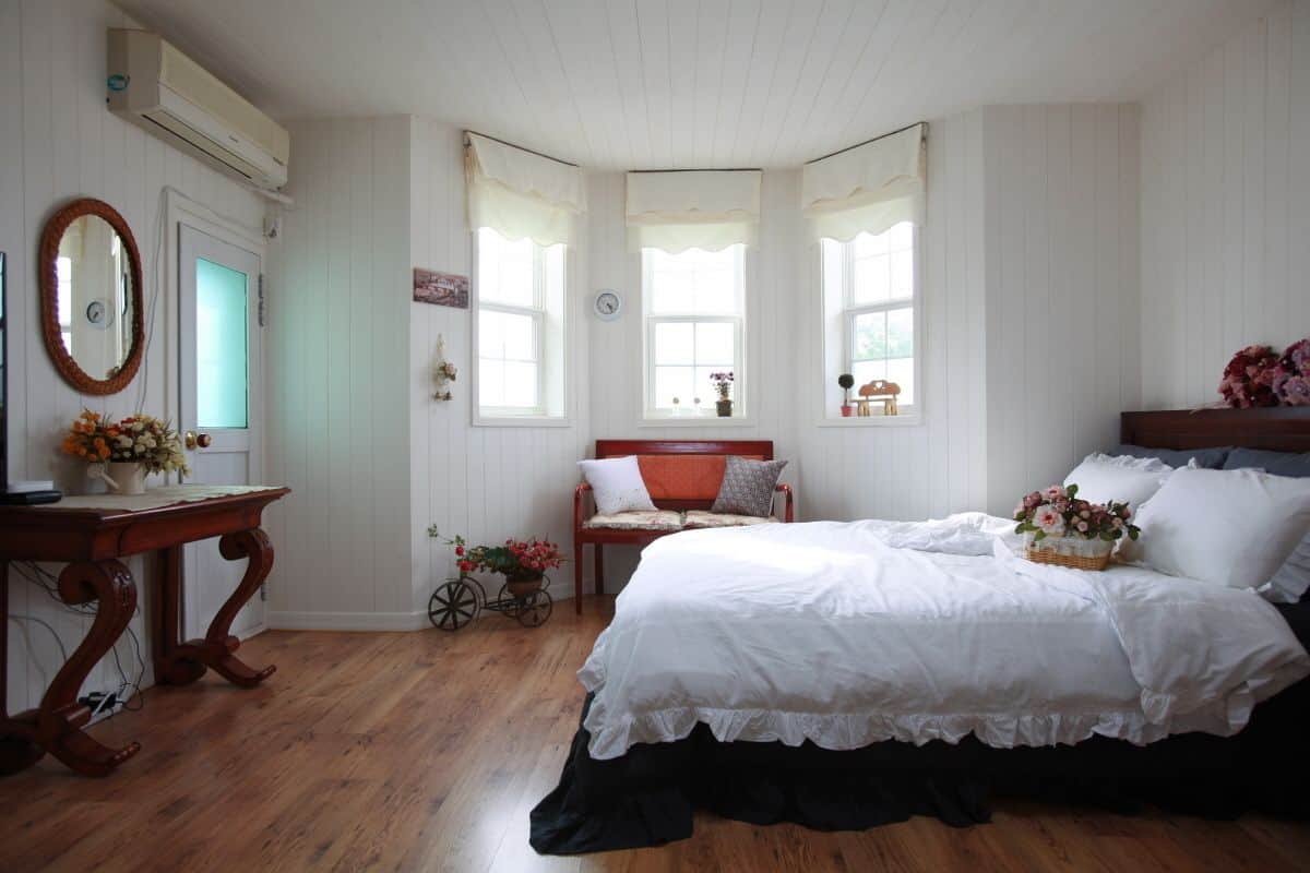Une chambre avec un plancher en bois, un lit avec une literie blanche, une commode en bois, un miroir et une fenêtre avec des rideaux.