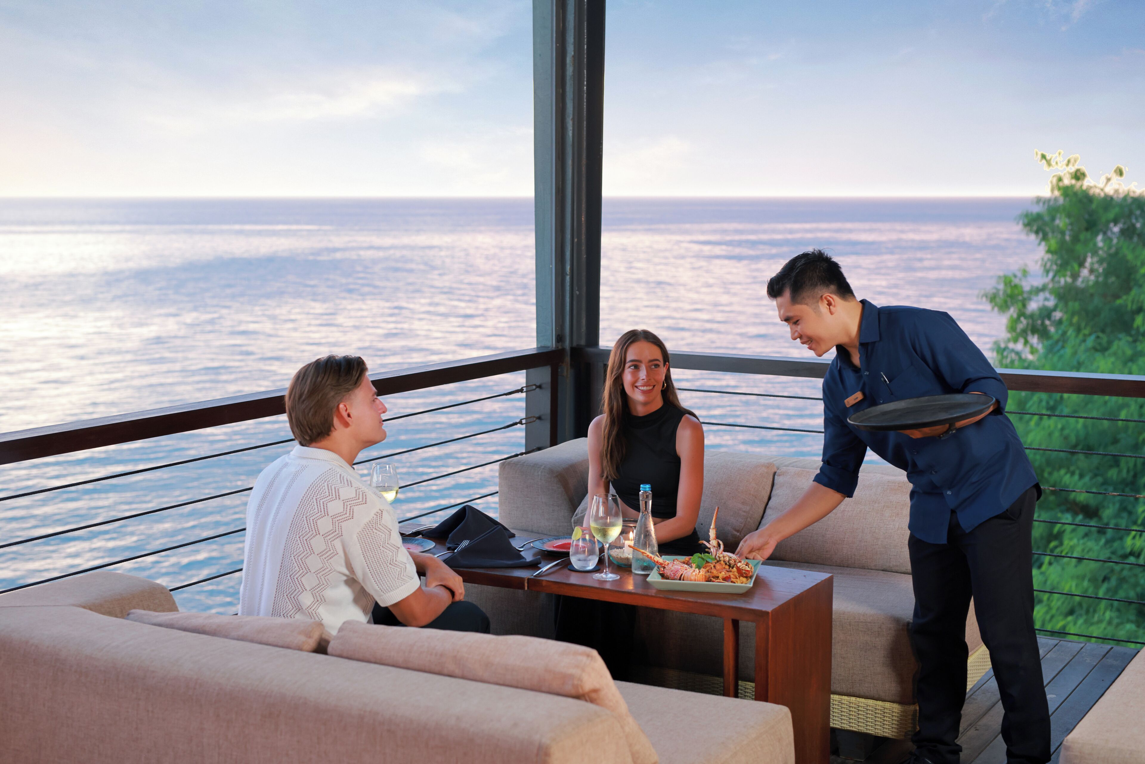 A dining area with a view of the sea, a table with a bottle, glasses, and a plate of food, and a waiter serving.