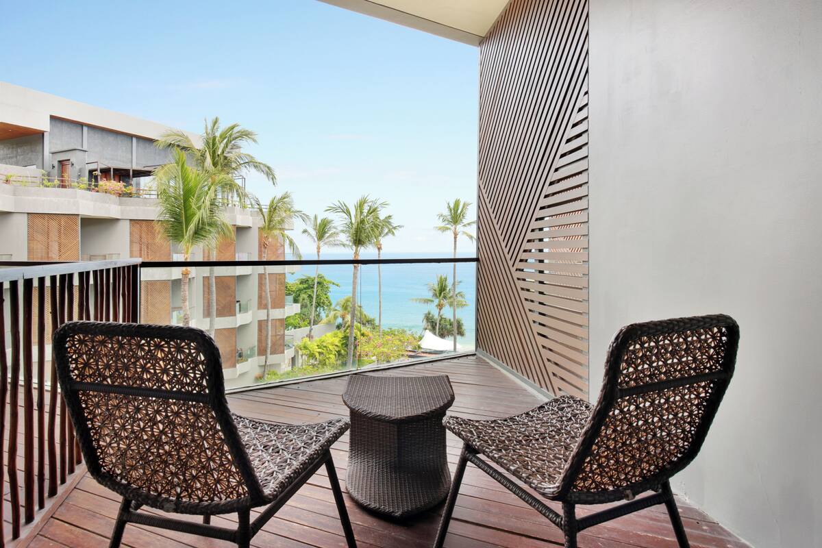A balcony with two wicker chairs, a small round table, and a view of the ocean and palm trees.