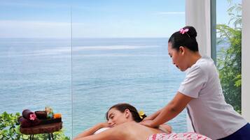 A spa setting with a massage table, a wooden side table with towels and a bowl, and a view of the ocean.
