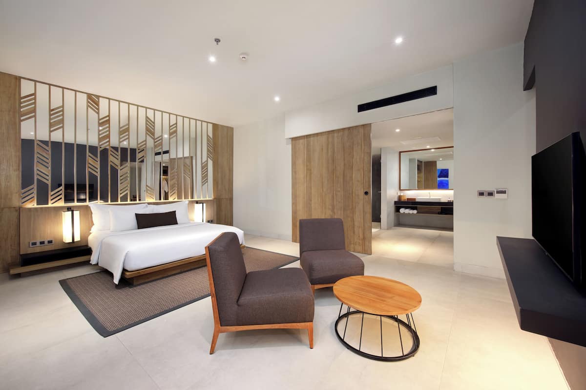 A modern hotel room with a bed, two armchairs, a round coffee table, a TV, and a large mirror.