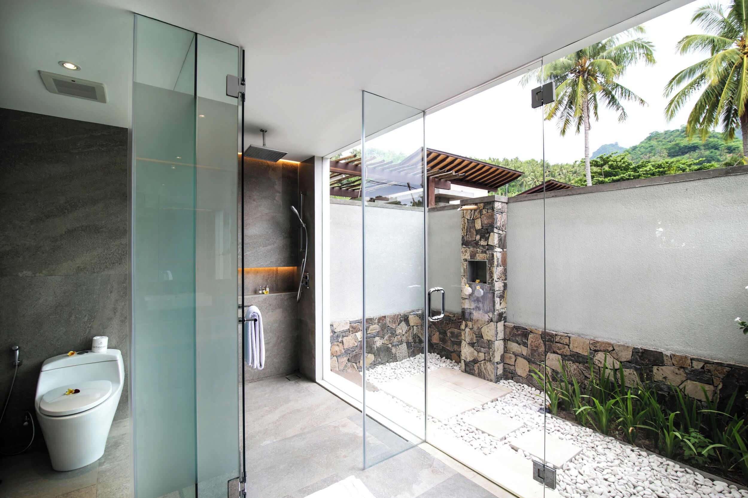 Un baño moderno con cabina de ducha de vidrio, pared de piedra y vistas a un paisaje tropical.