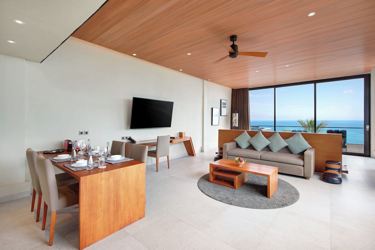 A modern living room with a dining area, a sofa, a flat-screen TV, and a large window overlooking the sea.