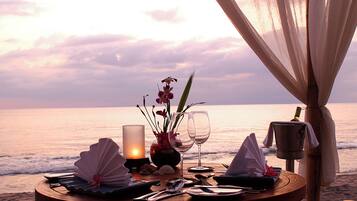 A romantic dinner setup on a beach with a table set for two, featuring a candle, wine glasses, and a decorative centerpiece.