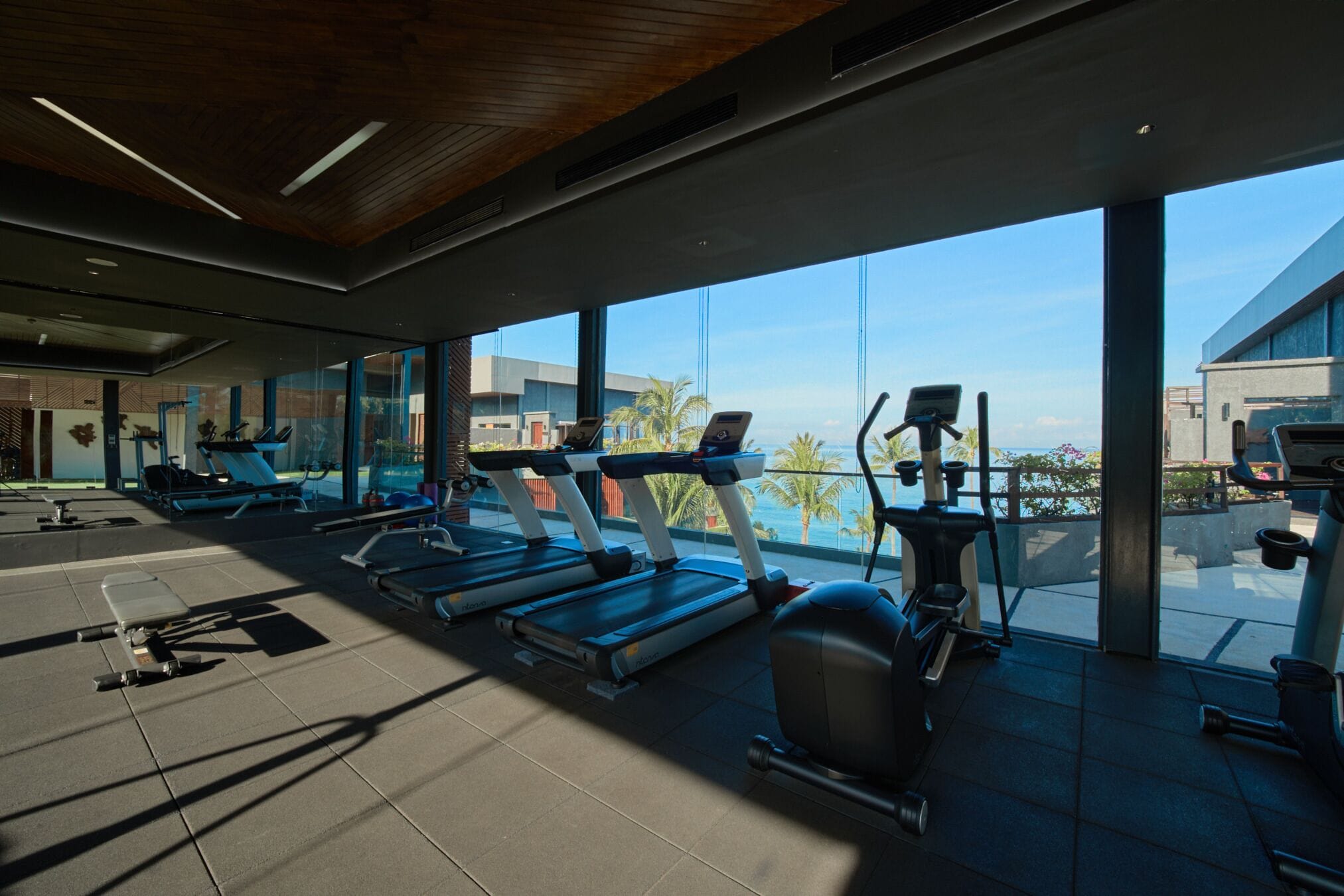 A modern gym with treadmills, elliptical machines, and exercise bikes, large windows with a view of palm trees and buildings.