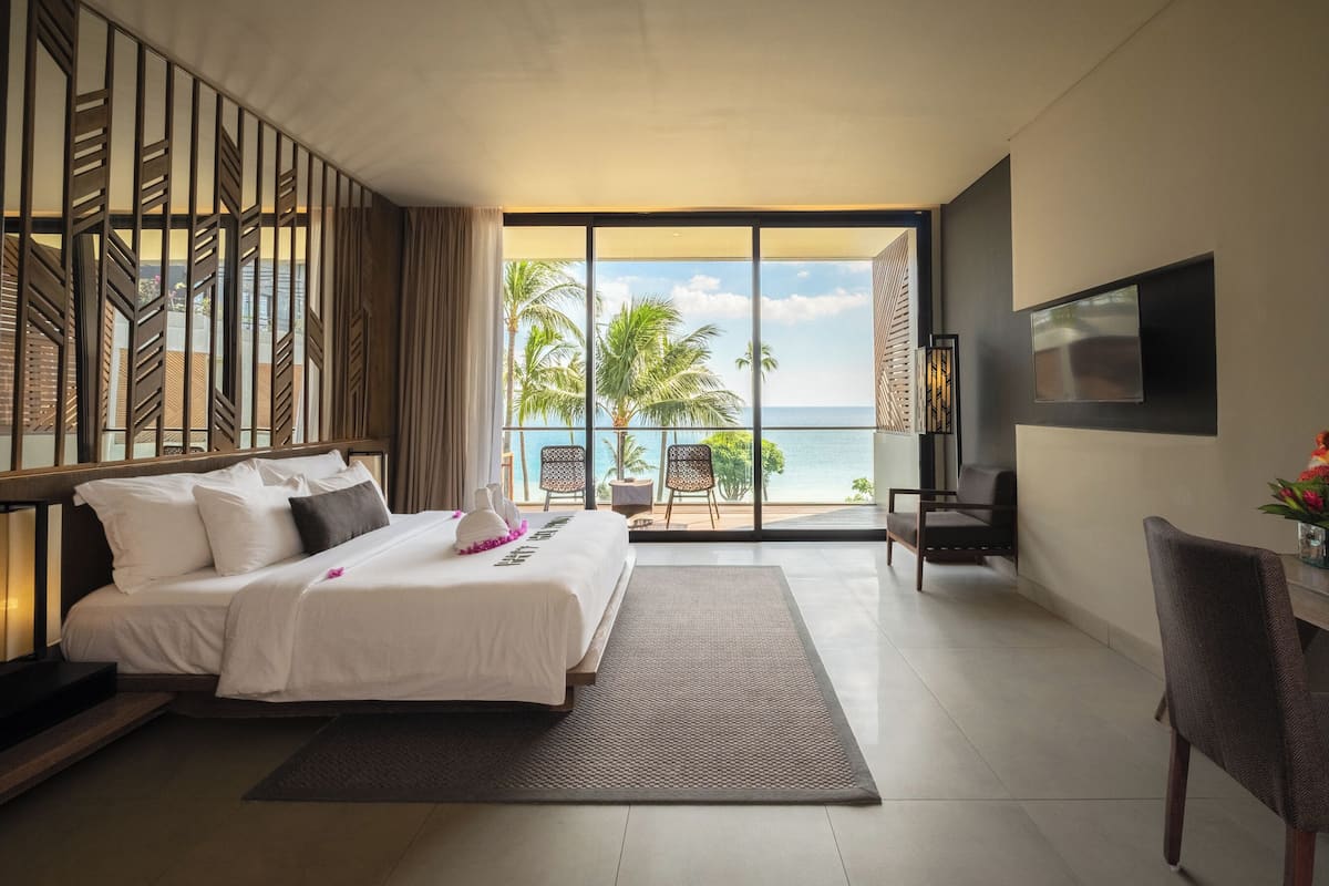 A hotel room with a bed, a desk, a chair, a TV, and a balcony with a view of the ocean and palm trees.