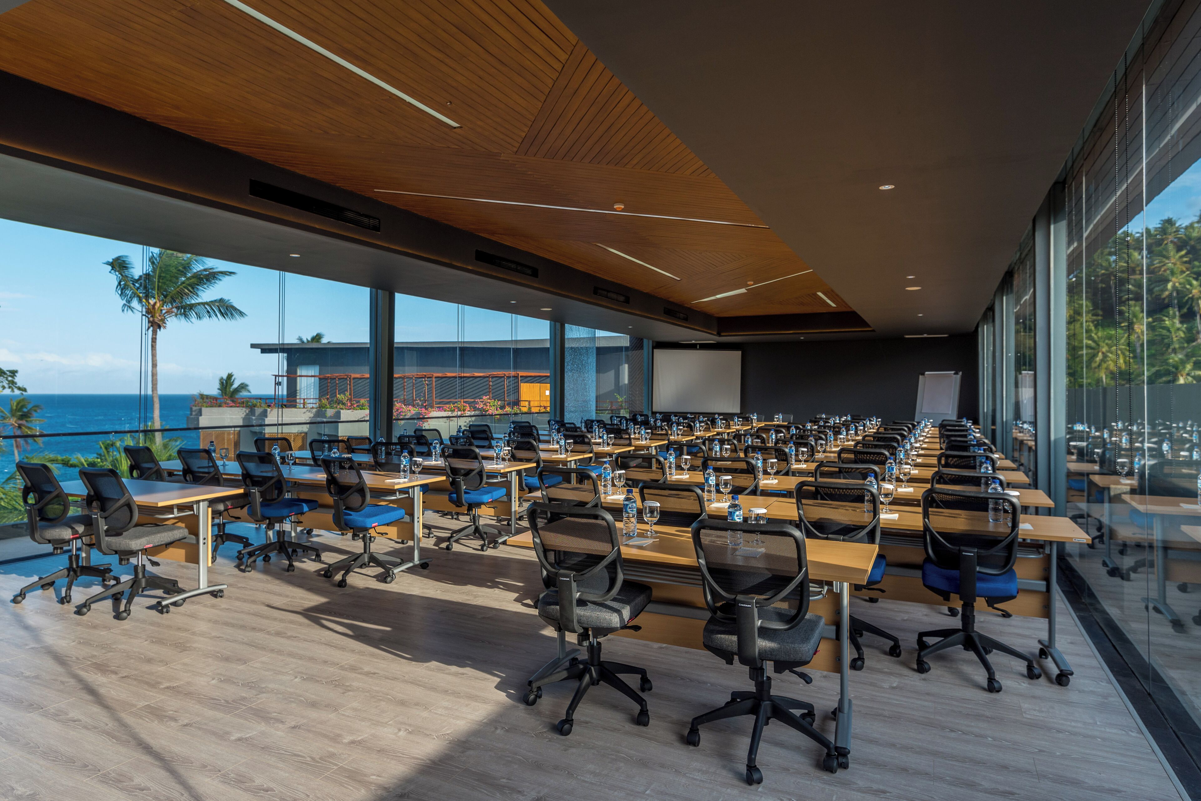 Un salón de conferencias con filas de mesas y sillas, ventanas amplias que dan a vistas del océano y palmeras, y un techo moderno de madera.