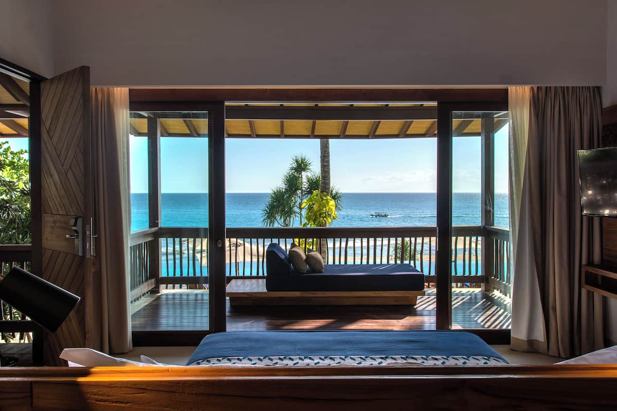 A room with a large window, a bed, a wooden desk, and a view of the ocean.
