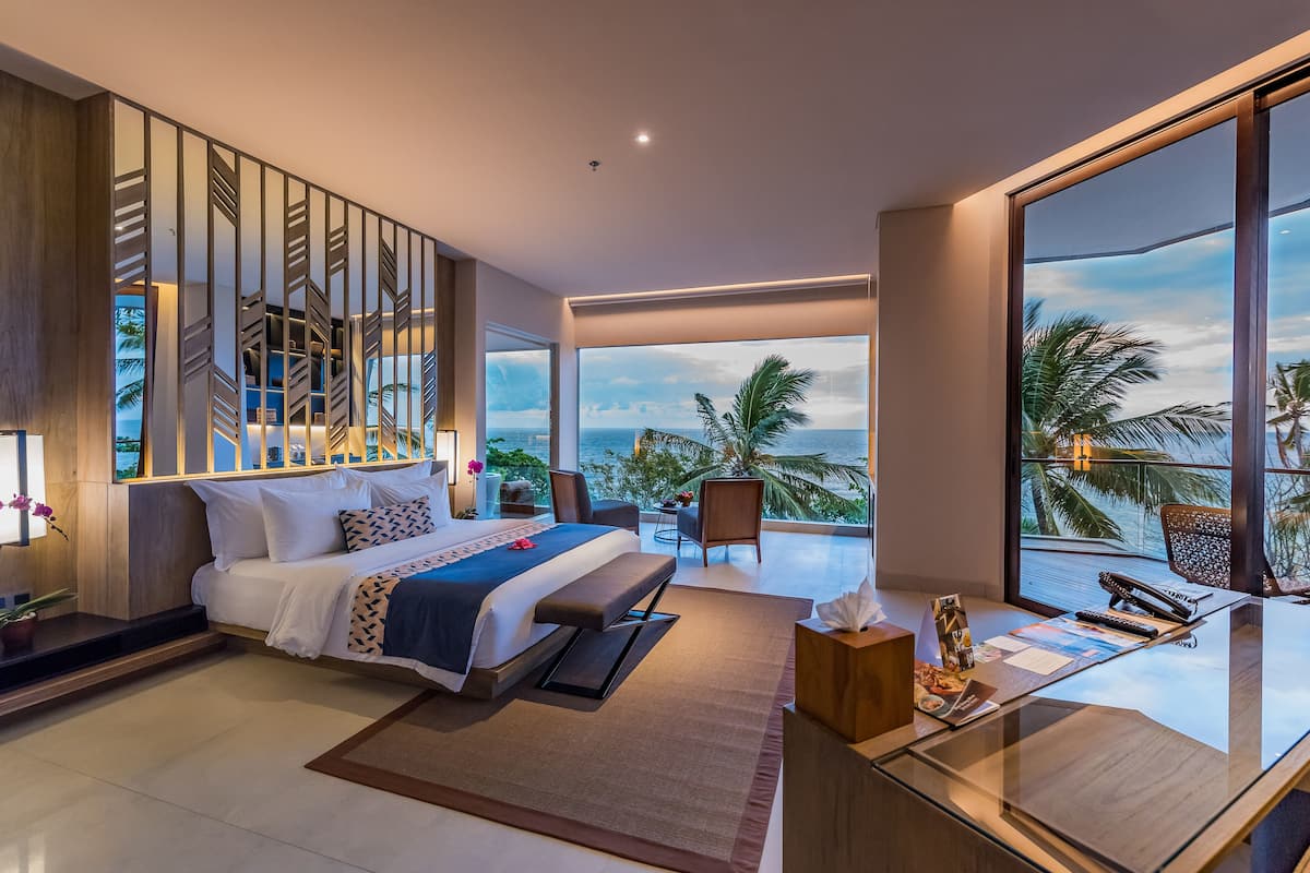 A modern hotel bedroom with a large bed, a desk, and a view of the ocean.