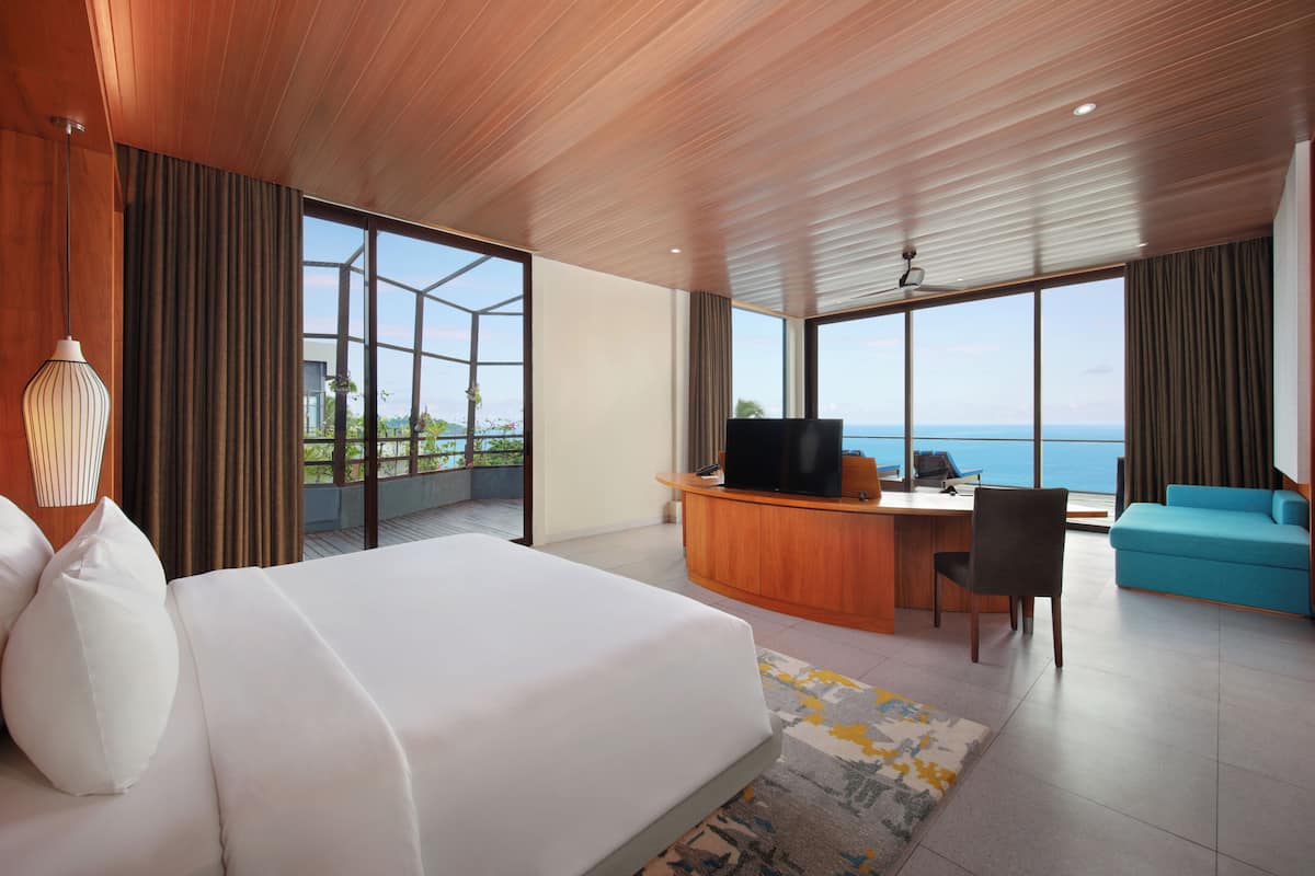 A modern hotel room with a large bed, a desk with a TV, a blue sofa, and a view of the ocean.