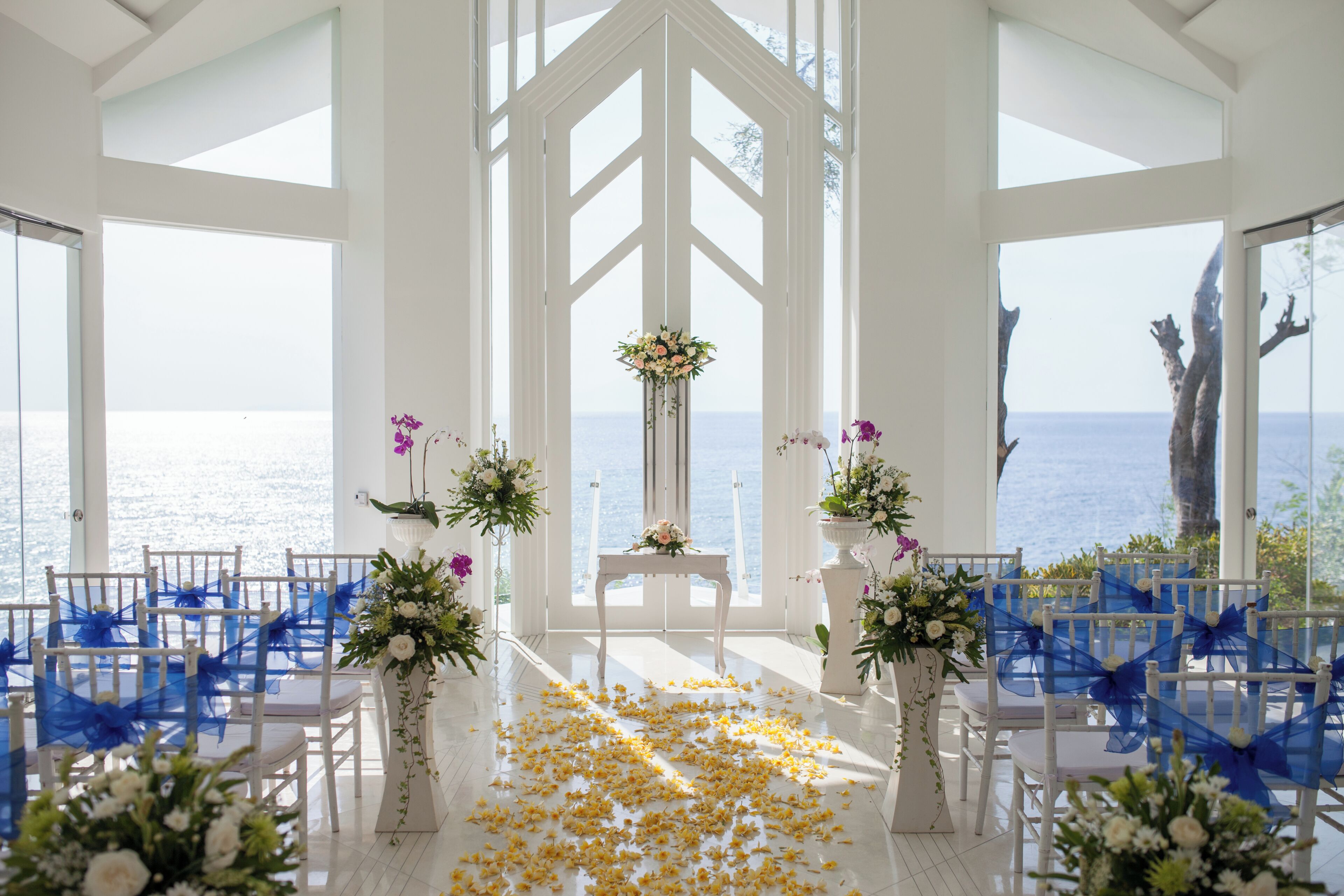 An indoor venue with white chairs, a central aisle, and floral arrangements.