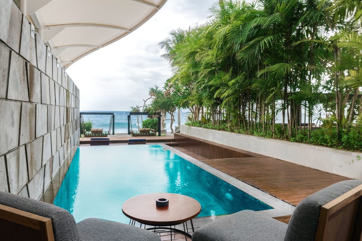 A swimming pool with a view of the ocean, surrounded by greenery and a covered seating area.