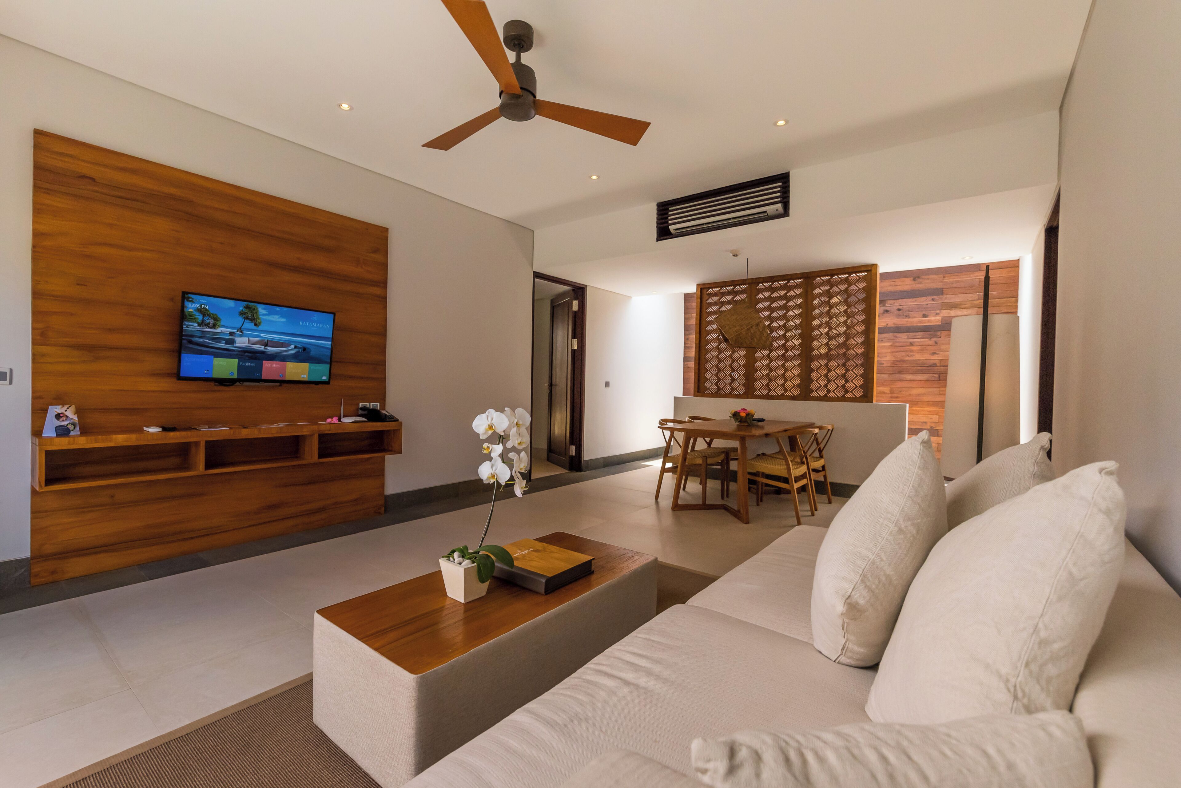 Uma sala de estar moderna com um móvel para entretenimento de madeira, uma TV de tela plana, um ventilador de teto e uma  área de jantar com mesa e cadeiras.