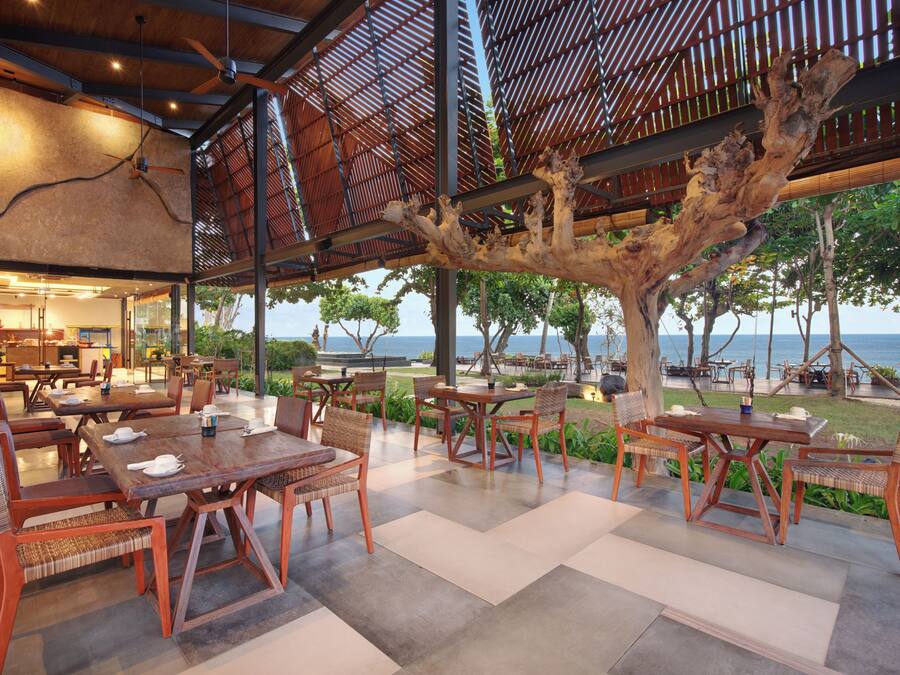 Un área de comedor al aire libre con mesas y sillas de madera, un gran árbol y vista al mar.