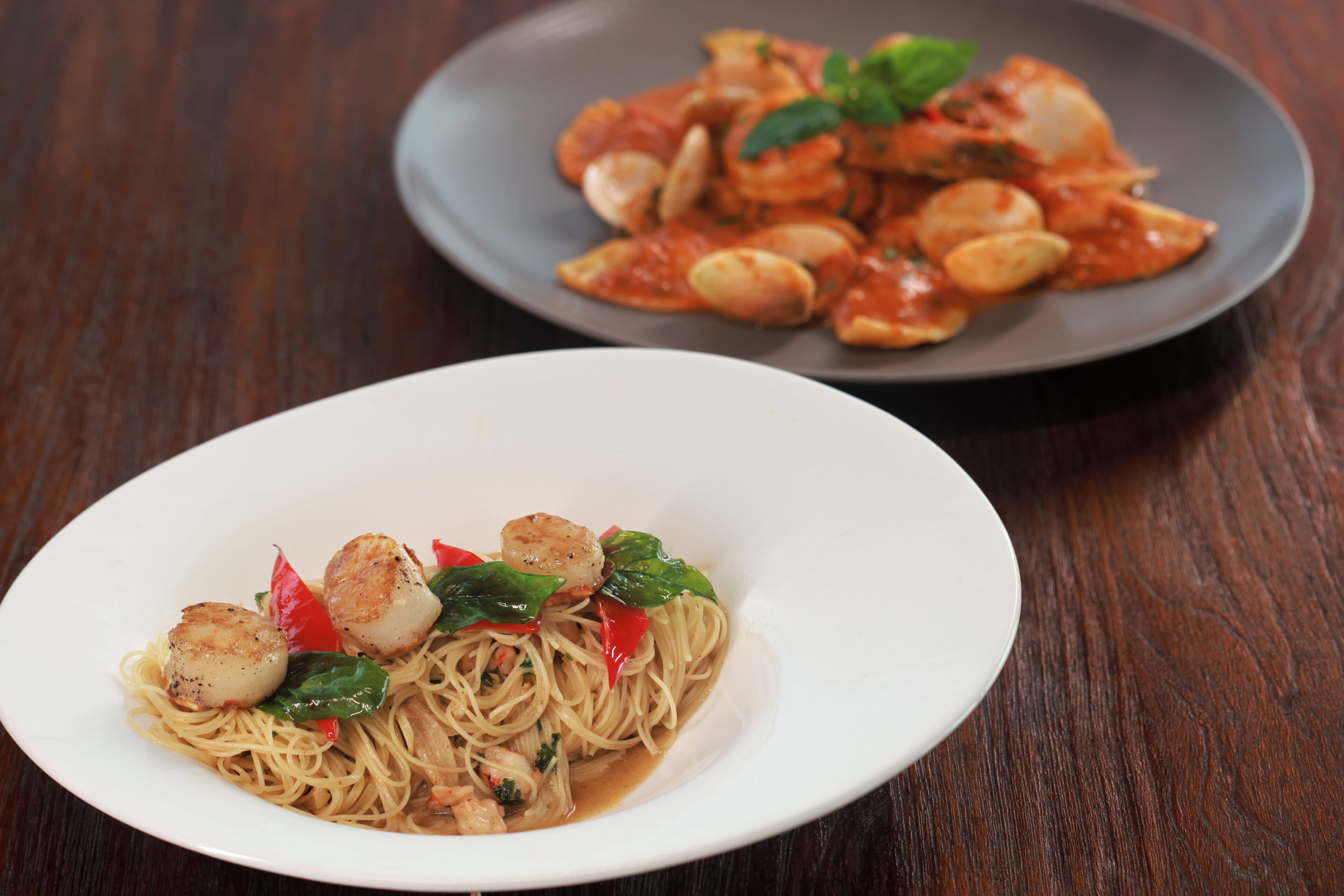 Two plates of pasta with vegetables and seafood on a wooden table.