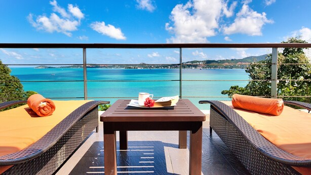 A balcony with wicker furniture, a table with a book and a cup, and a view of a turquoise sea and distant land. - Villa Seven Swifts (Koh Samui)