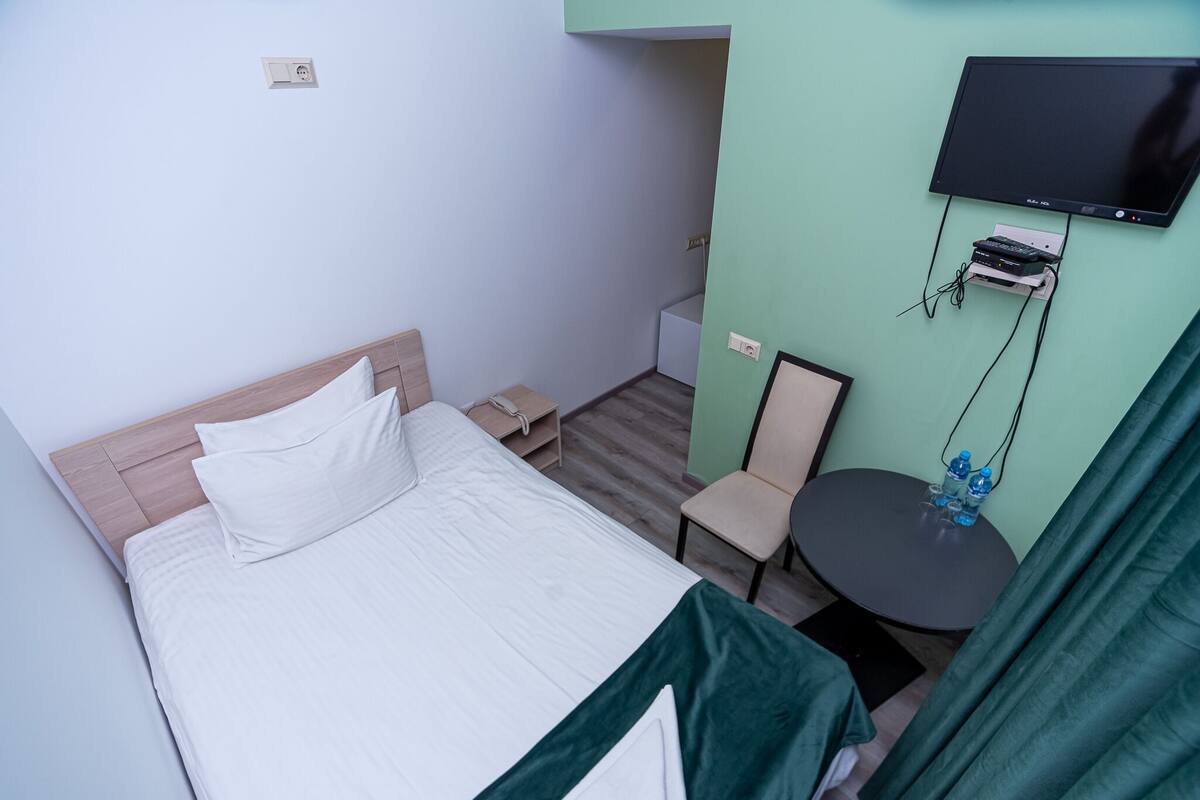Habitación de hotel con cama, una mesita con silla, televisor montado en la pared y toma de corriente.