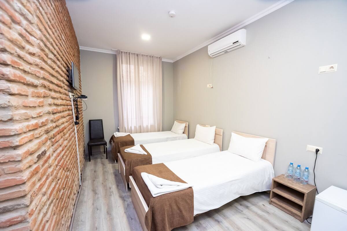 Habitación de hotel con tres camas, una pared de ladrillo, una ventana con cortinas, un equipo de aire acondicionado y una mesita de noche con botellas de agua.