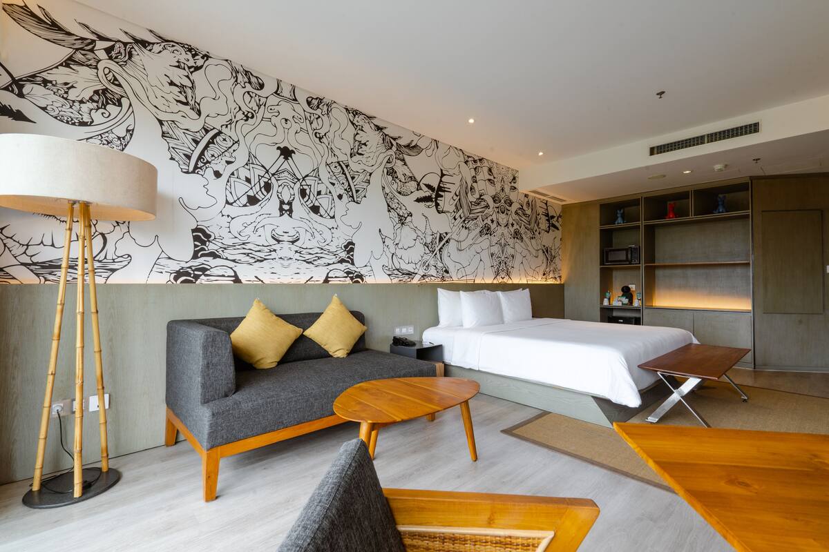 A modern hotel room with a large bed, a sofa with cushions, a round wooden coffee table, and a floor lamp.