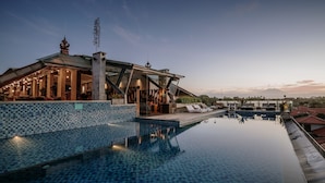 Una piscina infinita en la azotea con borde de azulejos, un área de estar cubierta con sillones de descanso y vistas a edificios lejanos.